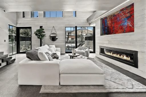 White modular luxury sectional sofa with oversized ottoman in a contemporary open-concept living room featuring a linear fireplace, modern artwork, and floor-to-ceiling windows.